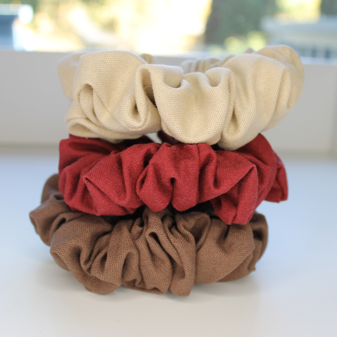 Three scrunchies in beige, red, and brown on a white surface with a blurred background.