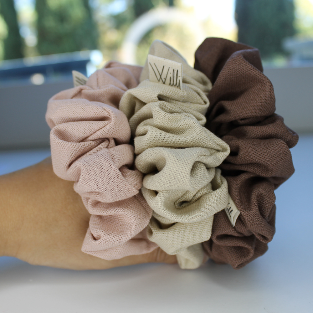 Set of four scrunchies in pink, beige, and brown on a white surface with blurred outdoor background.