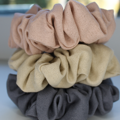 Three fabric scrunchies in beige, brown, and gray on a blurred natural background