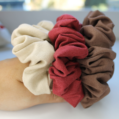 Three scrunchies in beige, red, and brown held by a hand on a blurred background