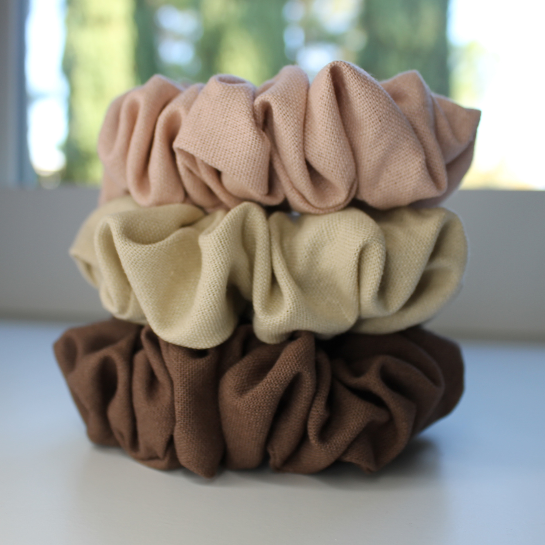 Stack of three scrunchies in brown, beige, and pink on a white surface with a blurred outdoor background.