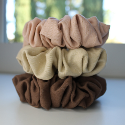 Stack of three scrunchies in brown, beige, and pink on a white surface with a blurred outdoor background.