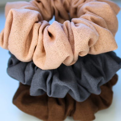 Three fabric scrunchies in pink, gray, and brown on a light background