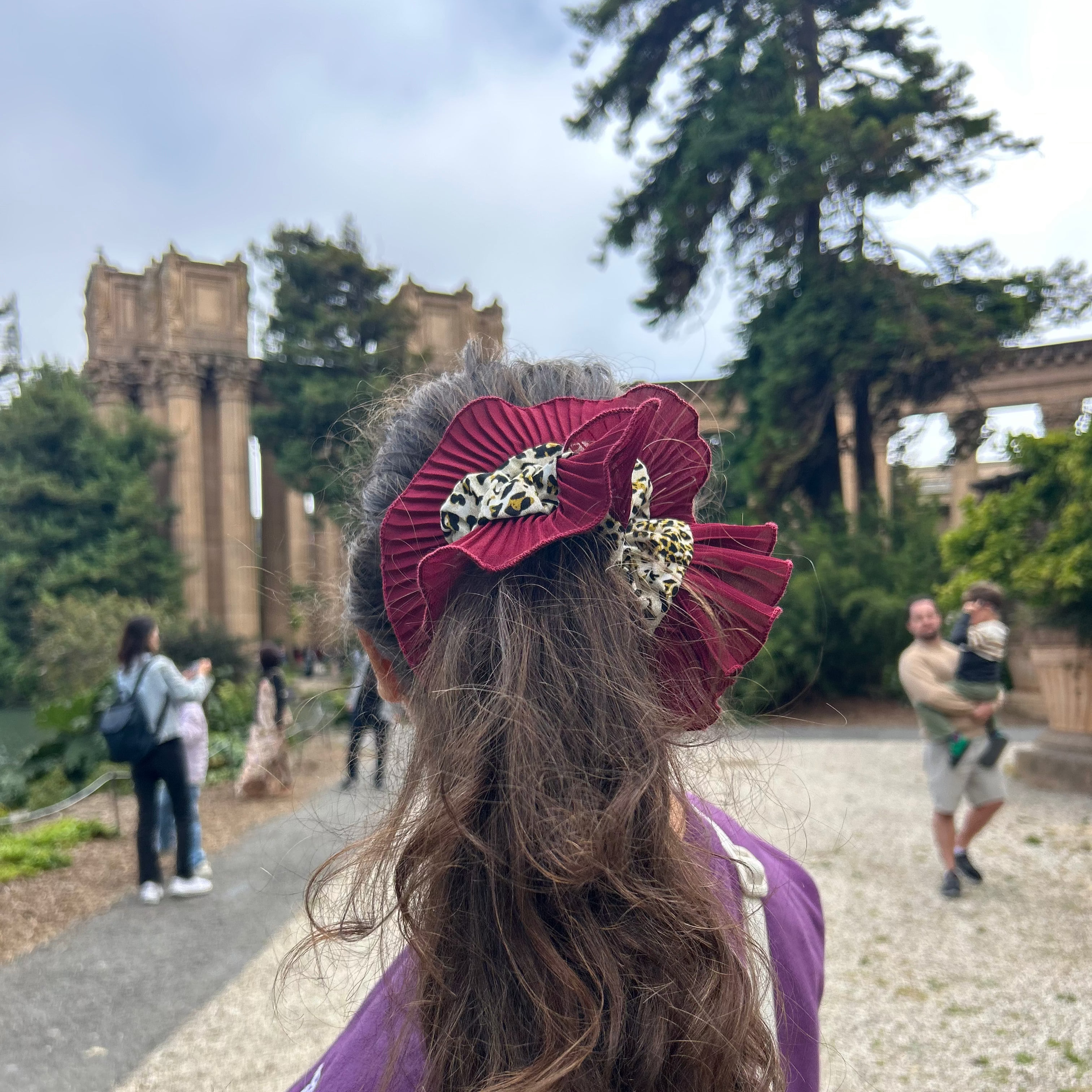 Person with a large decorative hair scrunchie outdoors, with trees and a building in the background.