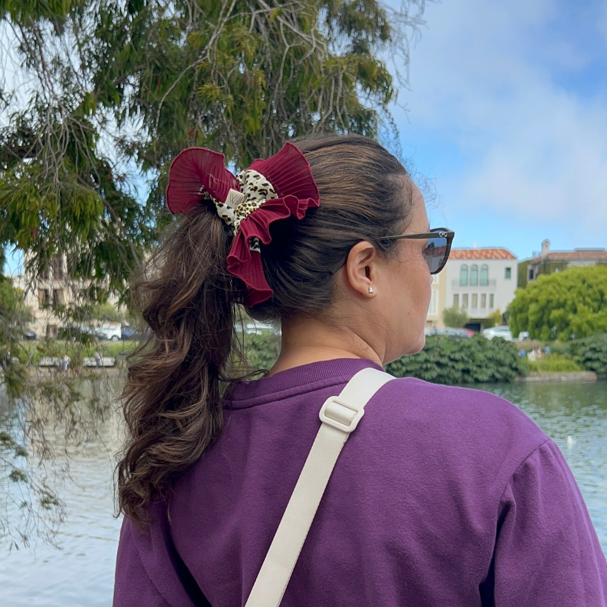 Person wearing a purple sweatshirt  and sun glasses standing by a lake with trees and buildings in the background using a large decorative red scrunchie