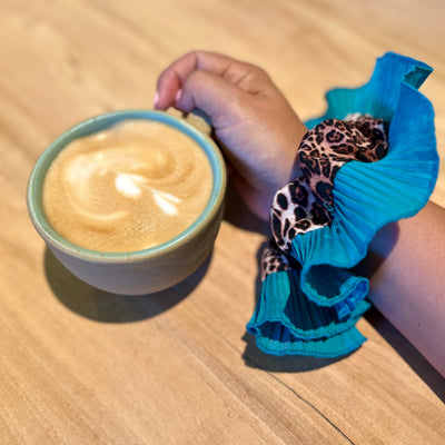 Hand holding a cup of coffee with a blue scrunchie and leopard print hair tie on a wooden surface